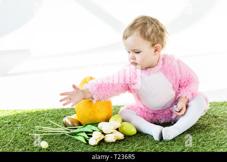 Adorable kid in rosa Flauschige Kostüm in der Nähe von Yellow Straußenei sitzen, bunte Hühnereier und Tulpen isoliert auf weißem Stockfoto