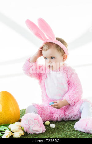 Adorable Kind in Funny Bunny Kostüm in der Nähe von Yellow Straußenei sitzen, bunte Ostereier und Tulpen isoliert auf weißem Stockfoto