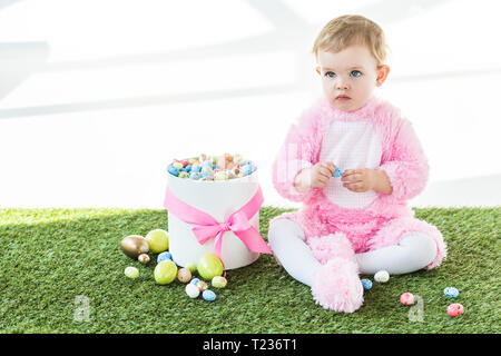 Adorable Baby in rosa Flauschige Kostüm sitzen auf grünem Gras in der Nähe der Box mit bunten Ostereier isoliert auf weißem Stockfoto