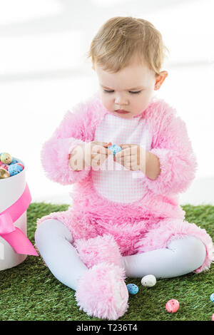 Cute kid in rosa Flauschige kostüm Holding blau Wachtelei beim Sitzen in der Nähe der Box mit Ostereier auf weißen isoliert Stockfoto