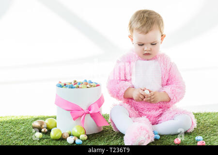 Cute Baby in rosa Flauschige Kostüm holding Wachtelei beim Sitzen in der Nähe der Box mit bunten Ostereier auf weißen isoliert Stockfoto