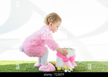 Adorable Kind in rosa Flauschige Kostüm steht in der Nähe der Box mit rosa Schleife und bunte Ostereier isoliert auf weißem Stockfoto