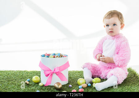 Cute Baby in rosa Flauschige Kostüm sitzt in der Nähe der Box mit bunten Ostereier isoliert auf weißem Stockfoto