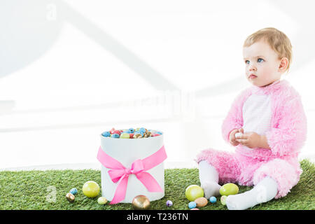 Adorable Kind in rosa Flauschige Kostüm sitzt in der Nähe der Box mit rosa Schleife und bunte Ostereier isoliert auf weißem Stockfoto