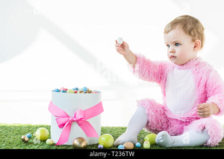 Adorable Kind in rosa Flauschige Kostüm holding Wachteleier in der ausgestreckten Hand beim Sitzen in der Nähe der Box mit bunten Ostereier isoliert auf weißem Stockfoto