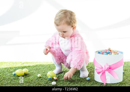 Adorable kid in rosa Flauschige Kostüm, bunte Eier aus grünem Gras in der Nähe von Geschenkbox mit rosa Schleife isoliert auf weißem Stockfoto