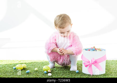 Cute Baby in rosa Flauschige Kostüm sitzen auf grünem Gras in der Nähe von Geschenkbox mit rosa Schleife und bunte Ostereier isoliert auf weißem Stockfoto