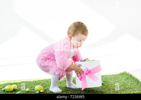 Adorable kid in rosa Flauschige Kostüm holding Geschenkkarton mit rosa Schleife und bunte Ostereier isoliert auf weißem Stockfoto