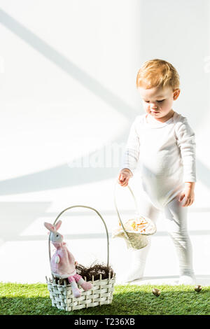 Adorable Baby in der Nähe von Stroh Korb mit dekorativen Kaninchen auf Weiß Stockfoto