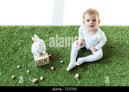 Adorable kid sitzen auf grünem Gras in der Nähe von Holz- Kalender mit 28. April Datum, dekorative Hase und bunte Ostereier isoliert auf weißem Stockfoto
