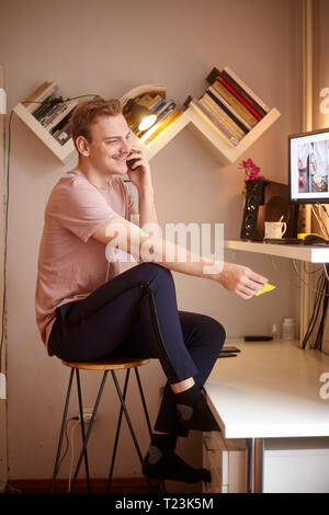 Ein junger Mann, in Barhocker sitzen, lächeln und sprechen über Telefon, zu Hause. Schreibtisch mit Computer Equipment vor ihm. Stockfoto