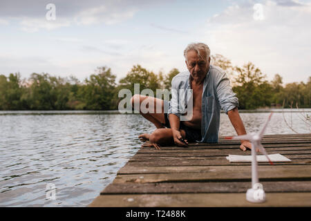 Älterer Mann sitzt auf Steg an einem See mit kleinen wind turbine Modell Stockfoto
