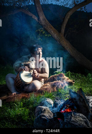 Caveman gekleidet in Wolf haut Drum spielt in der Nähe von Feuer im Wald Stockfoto