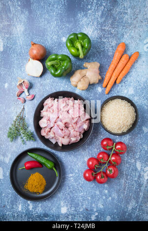 Blick von oben auf rohem Huhn und Reis in dunklen Schüsseln mit Wurzelgemüse auf der Arbeitsplatte in der Küche. Indredients für Afrikanische jollof Reis. Zutaten für Exoti Stockfoto