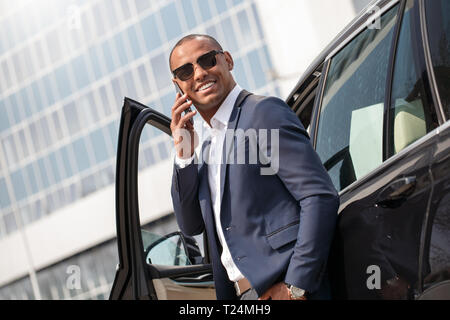 Junge afrikanische amerikanische Mann mit Sonnenbrille, lehnte sich auf dem Auto Reden auf dem Smartphone beiseite schaut lächelnd heiteres Seitenansicht Stockfoto