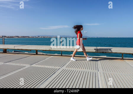 Junge Frau in rotem Kleid, unordentlichen Haaren und Telefon gehen durch den Betonpier, Valencia Hafen, Spanien Frau allein in rotem weiß Stockfoto