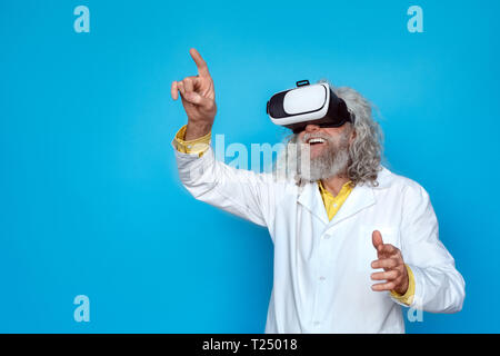 Alte langhaarigen Mann, der Doctor's Kleid und Virtual Reality headset Studio auf der blauen Wand in Bild zeigen isolierte lächelnd beiseite suchen Stockfoto