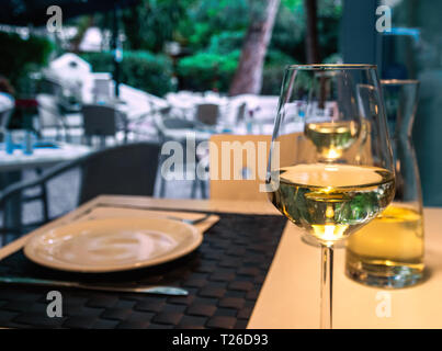 Ein Glas Weißwein, Karaffe, weißer Teller und Besteck auf einem Tisch in einem Straßencafé. Essen im Freien, gute Mahlzeiten, Wochenende. Selektiver Fokus Stockfoto