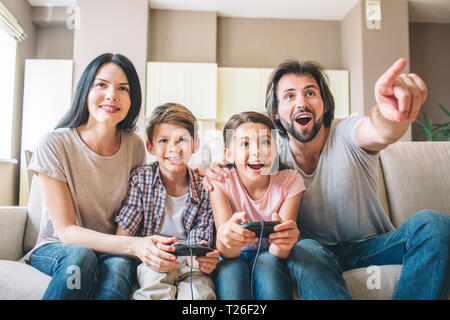 Die Eltern sitzen mit ihren Kindern auf dem Sofa. Kinder spielen das Spiel auf Video Spiel. Kerl ist nach vorne gerichtet und Schreien. Frau hilft zu spielen Stockfoto