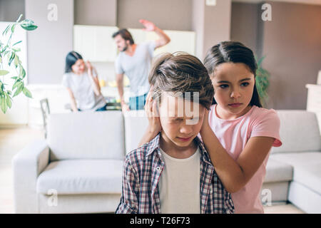 Mädchen steht hinter ihrem Bruder und hält seine Ohren mit den Händen geschlossen. Junge ist in Angst und Schrecken. Er hat die Augen geschlossen. Frau versucht, sich zu schützen. Kerl ist Stockfoto