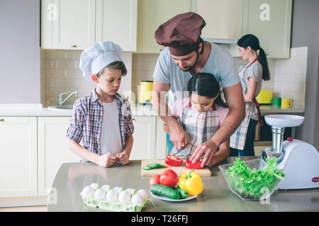 Vati lehrt Mädchen heiß Tomaten zu schneiden. Er hilft ihr. Junge sieht. Er ist konzentriert. Frau steht hinter ihnen, und Köche Stockfoto