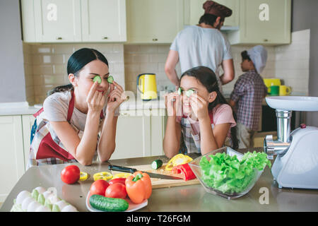 Moom und Tochter steht zusammen und lehnt sich an den Tisch. Sie spielen mit runden Stücke Gurke. Sie haben es auf die Augen. Jungen sind Arbeiten am Herd. Stockfoto