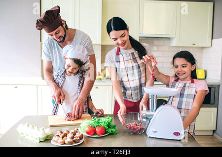 Vier Mitglieder der Familie zusammen. Kerl arbeitet mit seinem Sohn. Schneiden Sie Fleisch. Kleine Mädchen spielt mit dem Stück von ihm. Sie will es in zu setzen Stockfoto