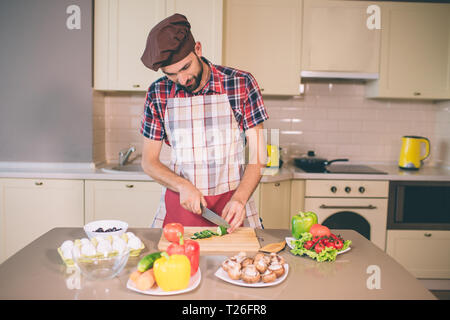 Vorsichtig Kochen steht und Gurke in kleine Stücke schneiden. Er tut es mit Messer. Kerl schaut. Er ist ernst und konzentriert Stockfoto