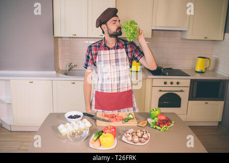 Bärtiger Mann steht und Gerüche Blätter von letuce. Er hält die Augen geschlossen. Mann ist ernst und konzentriert. Kerl steht in der Küche. Er bereitet Essen Stockfoto