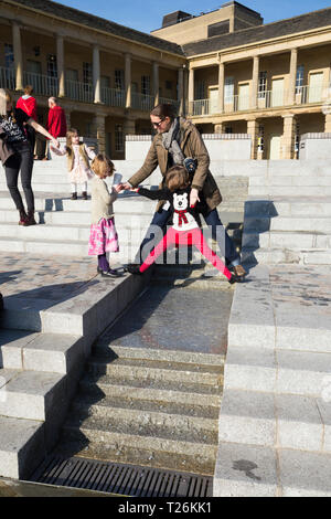 Kind spielt um das Wasserspiel & Besucher/Touristen sitzen und auf den Stufen im Viereck des wiederhergestellten Piece Hall entspannen. Sonnigen Tag/sun. Halifax. England UK. Der Tag war ein außergewöhnlich heißer Tag im Winter. Stockfoto