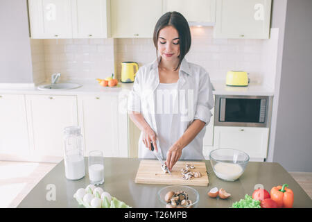 Ein Bild der dunkelhaarige Mädchen steht in der Küche und schneiden Pilze an Holz. Sie tut, vorsichtig. Mädchen konzentriert Stockfoto