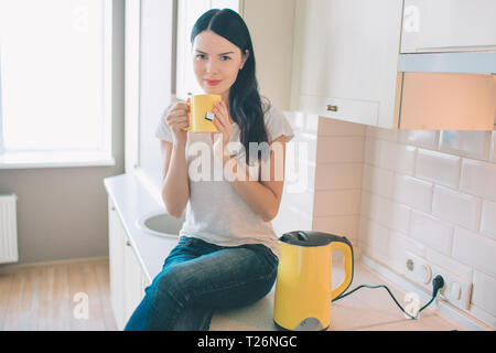 Nette Frau sitzt auf Bordsteinkanten. Sie posiert und ooks auf Kamera. Modell hält gelbe Schale in die Hände. Es gibt auch einen Wasserkocher neben ihr. Sie ist kühl und r Stockfoto