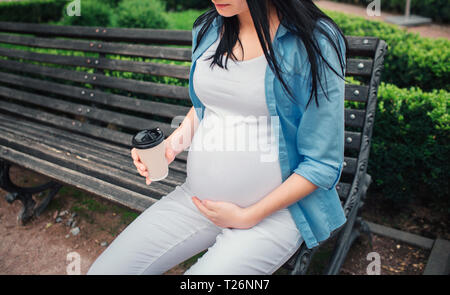 Portrait eines glücklichen schwarzen Haar und Stolz schwangere Frau in einer Stadt im Hintergrund. Das weibliche Modell sitzt auf einer Stadt Bank und trinken Kaffee o Stockfoto
