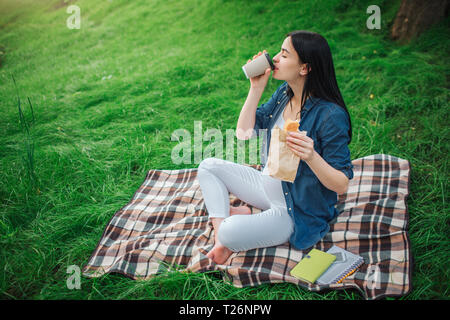 Portrait eines glücklichen schwarzen Haar und Stolz schwangere Frau in einer Stadt in den Park. Das weibliche Modell sitzt auf Gras und trinken Kaffee oder Tee und ea Stockfoto