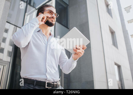 Positiver und fröhlicher junger Geschäftsmann steht bei hohen Gebäude und Posen. Er lächelt ein wenig. Kerl sucht gerade nach vorn. Er spricht am Telefon nd halten Piec Stockfoto