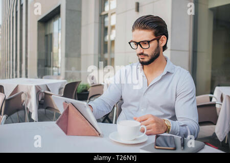 Konzentriert und stilvolle junge Mann sitzt am Tisch. Er arbeitet mit dem Tablet-PC. Junge Mann hält weiße Tasse Tee. Es gibt Notebooks mit Stift am Tisch. Er wor Stockfoto