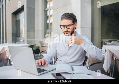 Konzentriert man sitzt am Tisch und funktioniert auf dem Laptop. Er halten Sie die Tasse Kaffee in der Hand. Es gibt Notebooks und Stift am Tisch geöffnet. Unternehmer arbeitet Stockfoto