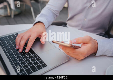 Schnittansicht der Hände des Menschen. Einer von ihnen ist das Liegen auf der Tastatur, während ein anderes Telefon in der Hand hält. Kerl benutzt es. Er sitzt am Tisch Stockfoto