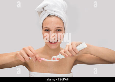 Positive sommersprossige junge Frau schaut auf Kamera und lächelt. Stellt Sie etwas Zahnpasta auf die Zahnbürste. Modell hat weiße Handtücher um ihren Körper und Haar. Ist Stockfoto