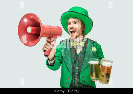 Glückliche junge Mann sprechen in Lautsprecher. Er halten es und Becher Bier in der Hand. Guy verschleiß St. Patrick. Auf grauem Hintergrund Stockfoto