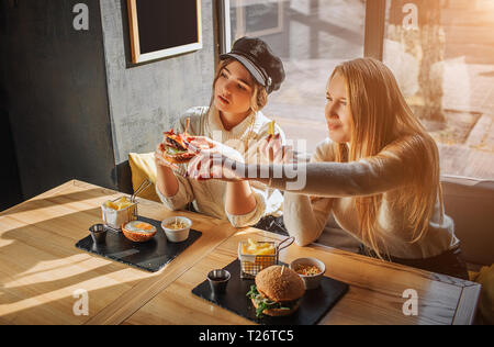 Zwei junge Frauen sitzen innen an Tabe. Modell auf der rechten Seite. Zweite halten Burger in der Hand. Sie freuen sich Stockfoto