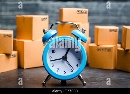 Kartons und einem blauen Wecker. Zeitpunkt der Lieferung. Begrenztes Angebot, Mangel an Waren auf Lager, Übertreibung und Verbraucher Fieber. Lieferung. conce Stockfoto