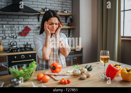 Junge kranke Frau hat Kopfschmerzen. Sie halten die Hände auf den Kopf. Sie sitzen in der Küche am Tisch. Frau schlecht fühlen. Gemüse auf dem Tisch liegend Stockfoto