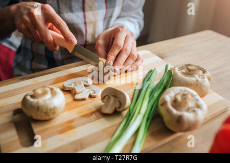 Nahaufnahme der Hand Mann mit Messer und Schneiden von Pilz. Frau, die ihm helfen, mit Kochen. Sie sitzen am Tisch. Grüne Zwiebeln und Pilze sind auf Tisch Schreibtisch Stockfoto