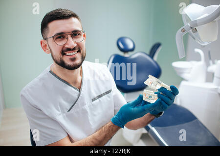 Schöne positive younng Zahnarzt in der Zahnheilkunde. Er halten künstliche Kiefer mit Zähnen und lächeln in die Kamera. Professionelle Arzt. Stockfoto