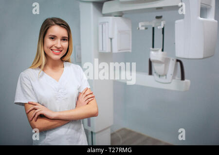 Nette junge weibliche Zahnarzt stehen in x-ray Zimmer und vor der Kamera posieren. Sie lächeln und die Hände gekreuzt. Professionell. Stockfoto