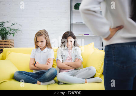 Traurig und wütend Teenager sitzen auf dem Sofa im Zimmer und nach unten schauen. Halten Sie Hände und Beine überkreuzt. Junge Frau stand vor ihnen. Sie halten die Hände an der Hüfte Stockfoto