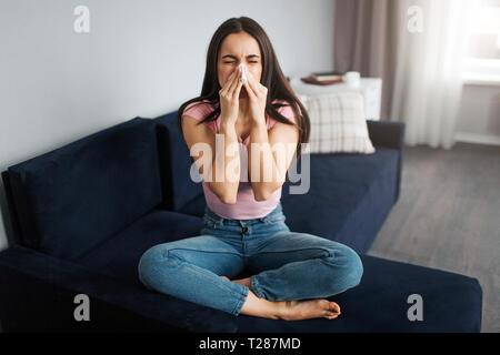 Bild der jungen Frau sitzen auf dem Sofa und Niesen in Weiß Serviette. Sie ist krank. Modell leiden. Sie fühlt sich schlecht Stockfoto