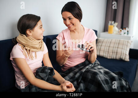 Fröhliche junge Frau und Mädchen sitzen zusammen auf der Couch und Lächeln. Sie schauen sich an. Mutter gießen Sirup in Löffel Stockfoto