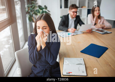 Kranke junge Frau saparately am Tisch im Konferenzraum sitzen und Niesen. Sie halten weiß Serviette. Ihre Kollegen arbeiten hinter Stockfoto